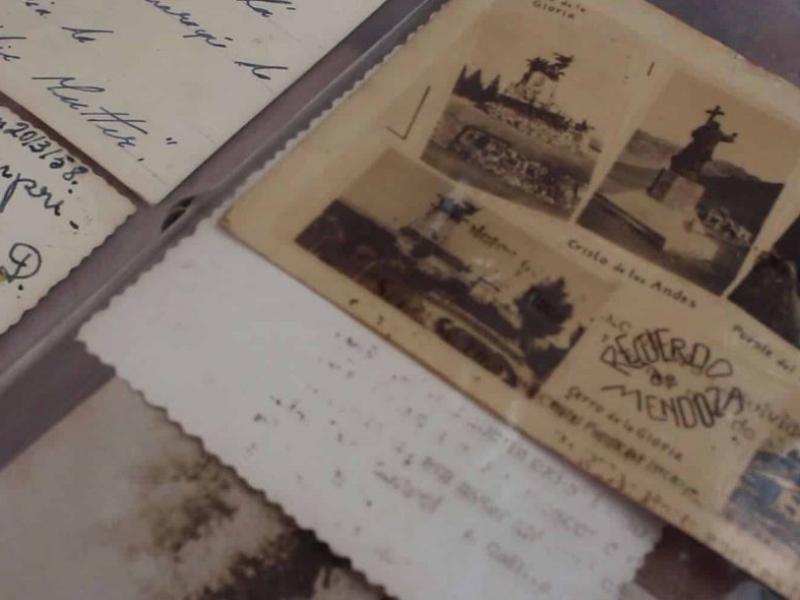Fotografia em close de documentos e imagens antigas sobre uma superfície. Em destaque, um cartão postal envelhecido com pequenas fotografias e a inscrição “Recuerdo de Mendoza”. Ao redor, há papéis com bordas desgastadas e trechos de escrita à mão, sugerindo correspondências antigas. A composição e os tons amarelados transmitem sensação de memória, arquivo e passagem do tempo.