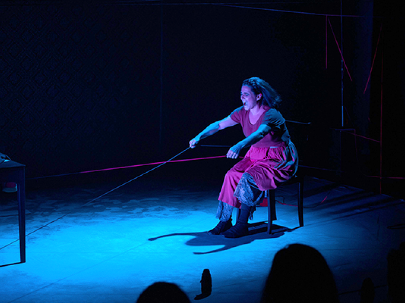Cena da peça Filipa mostra uma mulher sentada em uma cadeira, em palco escuro iluminado por luz azul. Ela veste vestido vermelho e puxa com força um fio ou corda esticada à sua frente, com expressão intensa e concentrada. O cenário é minimalista, com poucos objetos, e linhas finas coloridas atravessam o espaço, criando tensão visual e reforçando o clima dramático da cena.