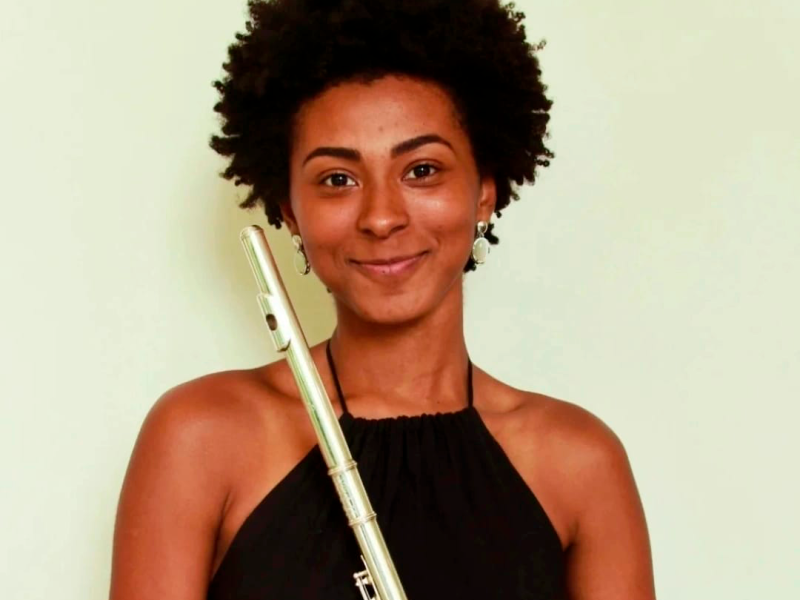 Retrato da Isabella Passos em fundo neutro claro. Ela sorri suavemente para a câmera, com cabelo curto e crespo. Veste blusa preta de alças e segura uma flauta transversal na vertical, próxima ao rosto. A iluminação é suave, destacando sua expressão acolhedora e a presença do instrumento.