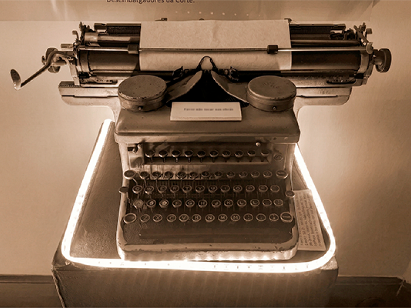 Máquina de escrever antiga em exposição. O objeto está sobre um pedestal iluminado, com papel encaixado no rolo e teclas circulares visíveis. A iluminação destaca os detalhes metálicos e o desgaste do equipamento, sugerindo valor histórico. Ao fundo, há uma parede neutra com pequena placa informativa acima.