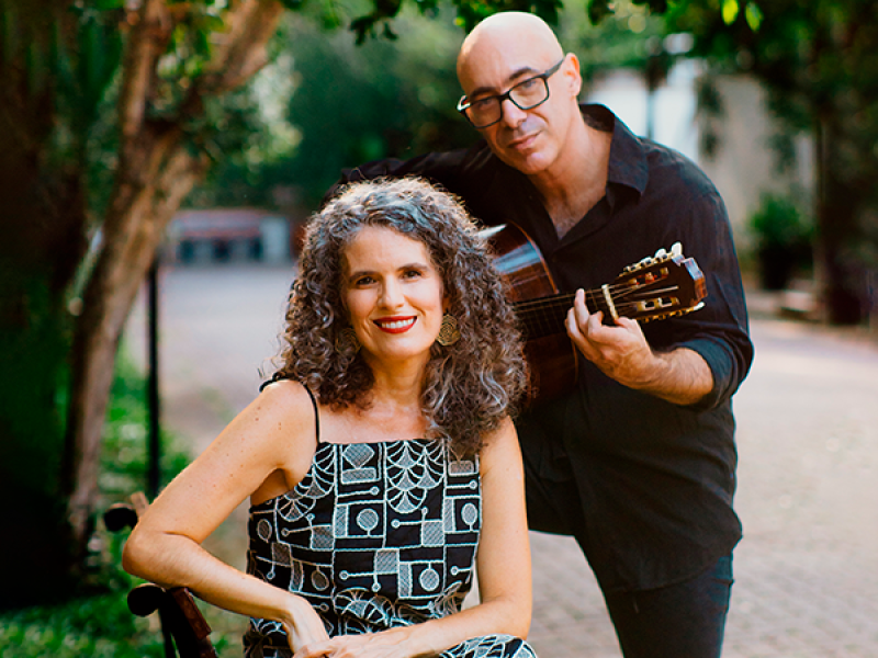 Fotografia de retrato ao ar livre da cantora e violonista Anna Paes e do violonista e cavaquinista Carlos Chaves. Em primeiro plano, Anna Paes, mulher de pele clara com cabelos cacheados grisalhos, está sentada em um banco de praça, sorrindo para a câmera, usando vestido estampado. Ao lado e ligeiramente atrás, Carlos Chaves, homem de pele clara, careca e com óculos, veste roupa escura e segura um violão, com postura de quem toca. O fundo mostra árvores e um caminho, criando um ambiente natural e acolhedor.