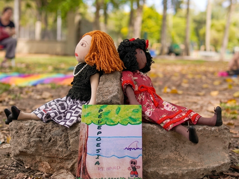 A imagem mostra um parque com árvores e folhas secas espalhadas pelo chão. Em primeiro plano, de frente para a câmera, há um livro com título PÉDEPOESIA apoiado em duas pedras, sobre o chão. Há duas bonecas de pano sentadas nas pedras, de costas uma para a outra. Uma das bonecas tem cabelos escuros e usa vestido vermelho; a outra tem cabelos claros e veste roupas em tons neutros. Ao fundo, há vegetação e um caminho sombreado, criando um ambiente natural e tranquilo.