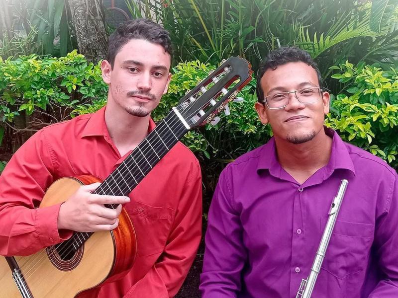 Retrato dos dois músicos do Duo Prevatto & Do Carmo ao ar livre, diante de folhagens. À esquerda, Tacio Prevatto segura um violão clássico, veste camisa vermelha e olha para a câmera com expressão tranquila. À direita, Erick do Carmo segura uma flauta transversal, usa camisa roxa e óculos, sorrindo levemente. A cena tem luz natural e clima informal, destacando a dupla e seus instrumentos.