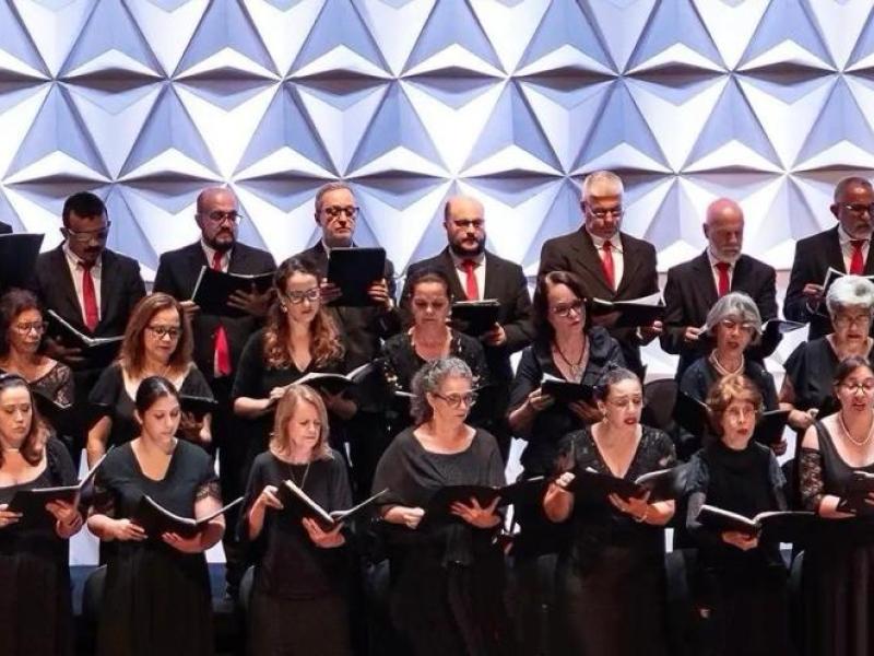 Apresentação de coral em palco mostra grupo numeroso de cantores organizados em fileiras. Todos vestem roupas formais em tons escuros, com alguns homens usando gravatas vermelhas. Cada integrante segura uma partitura e canta com expressão concentrada. Ao fundo, um painel geométrico branco cria textura visual. A iluminação é clara e uniforme, destacando o conjunto e a harmonia da apresentação.