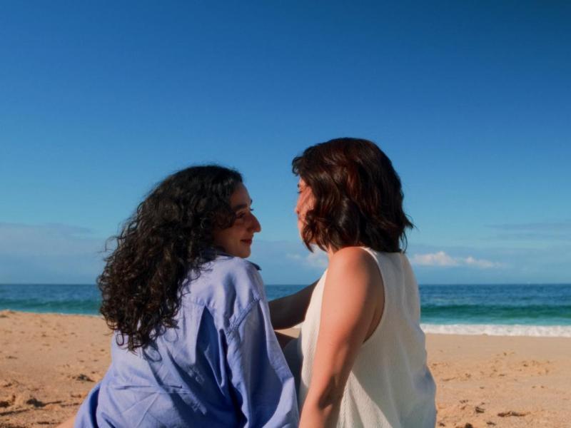 Cena do filme Azul mostra duas mulheres sentadas na areia de uma praia, próximas ao mar. Elas estão de perfil, voltadas uma para a outra, com expressões suaves e íntimas. Uma veste camisa azul e tem cabelos longos e cacheados; a outra usa blusa clara e tem cabelo curto. Ao fundo, o mar azul e o céu aberto criam um clima tranquilo e contemplativo.