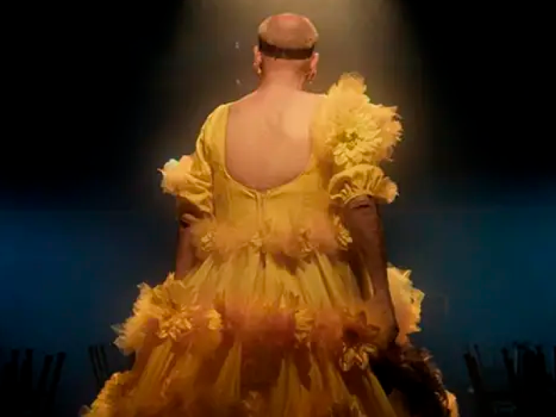 Cena do filme “Divinas Divas” mostra artista transformista de costas no palco, sob foco de luz dourada, usando vestido amarelo exuberante com flores aplicadas e mangas bufantes. O fundo escuro ressalta a dramaticidade da apresentação e a estética do espetáculo.