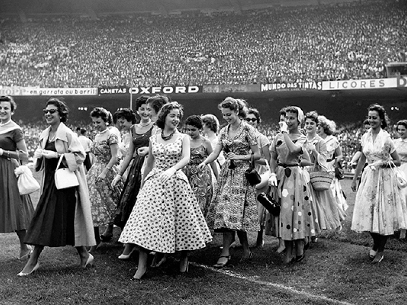 Imagem em preto e branco do documentário “Um filme à moda Bangu”. Um grupo de mulheres com vestidos rodados e estampados típicos dos anos 1950 caminha pelo gramado de um estádio lotado. Algumas sorriem e conversam entre si, enquanto a multidão aparece ao fundo nas arquibancadas. A cena remete ao universo da moda, do cotidiano e da cultura popular brasileira da época.