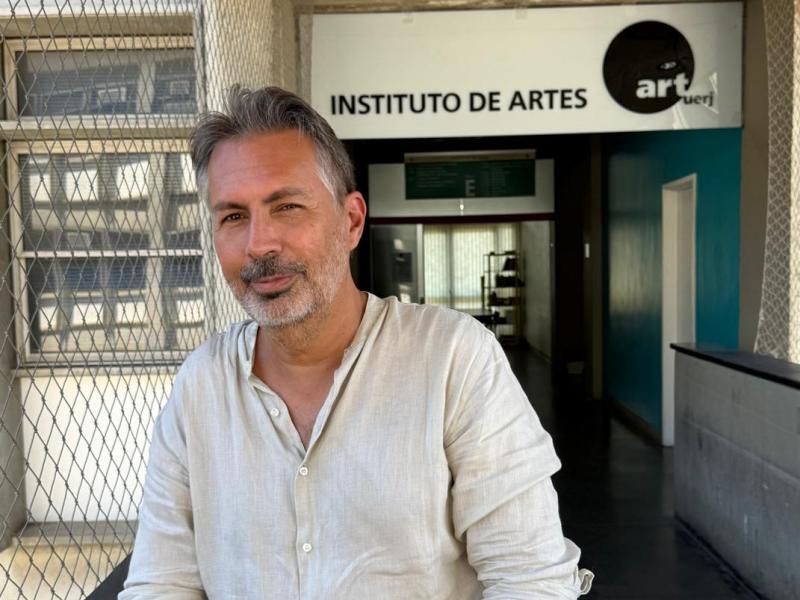 Retrato do professor Erick Felinto em área externa do Instituto de Artes da UERJ. Ele aparece em primeiro plano, com cabelos grisalhos e barba curta, usando camisa clara de linho e olhando para a câmera com leve sorriso. Ao fundo, vê-se a entrada do instituto, com a placa “Instituto de Artes” e parte do corredor interno do prédio.