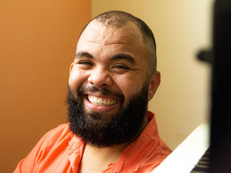 Retrato do pianista Thalyson Rodrigues sorrindo para a câmera. Ele aparece em primeiro plano, com barba cheia e camisa em tom alaranjado. À direita da imagem, parte desfocada de um piano sugere o contexto musical do ambiente. O fundo em tons quentes reforça a atmosfera acolhedora da cena.
