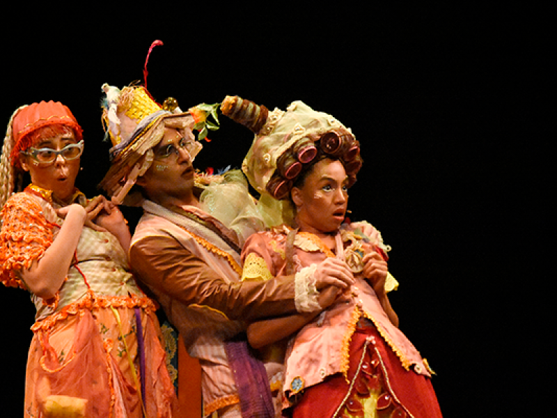 Cena da peça teatral “Era Uma Vez um Tirano”. Três atores aparecem juntos no palco, usando figurinos coloridos e extravagantes em tons de laranja, rosa e dourado, com maquiagem e acessórios exagerados. As personagens fazem expressões de surpresa e comicidade, inclinando os corpos para trás em movimento teatral. O fundo escuro destaca o clima lúdico e fantasioso da encenação.