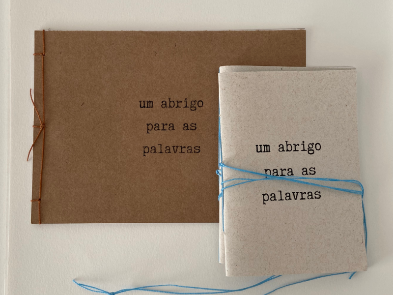 Fotografia de dois cadernos artesanais com capas em papel reciclado, posicionados sobre fundo claro. O maior, ao fundo, tem capa marrom e costura lateral com linha laranja. O menor, à frente, possui capa clara amarrada com barbante azul. Ambos trazem o título “um abrigo para as palavras” impresso em letras pretas, em composição simples e delicada.