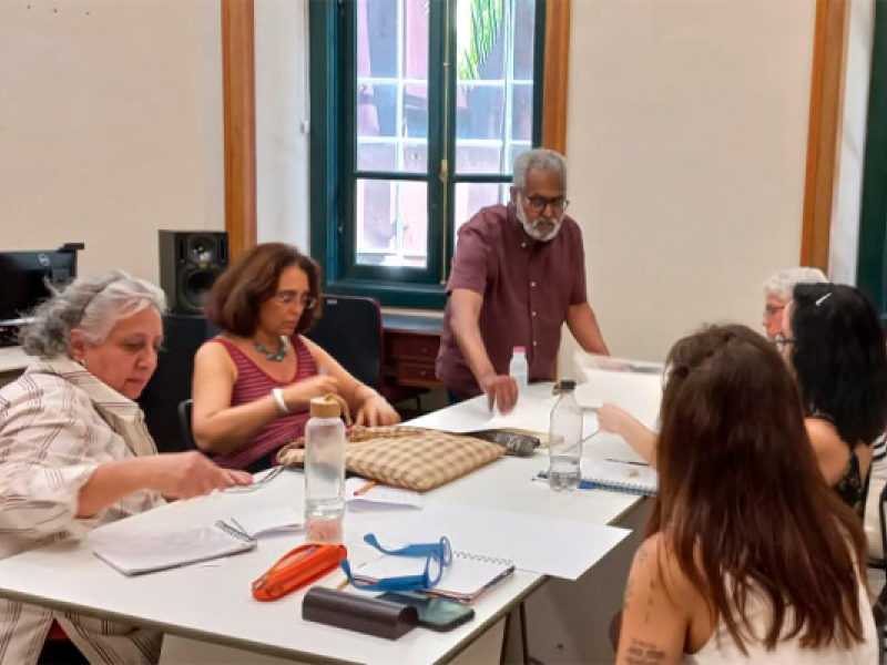 Grupo de pessoas reunidas em torno de uma mesa durante atividade ou oficina em sala interna. Um homem de barba grisalha está em pé, distribuindo folhas de papel, enquanto outras pessoas sentadas organizam materiais e fazem anotações. Sobre a mesa há cadernos, garrafas de água, estojos e papéis. Ao fundo, uma janela alta com moldura verde ilumina o ambiente de trabalho coletivo.