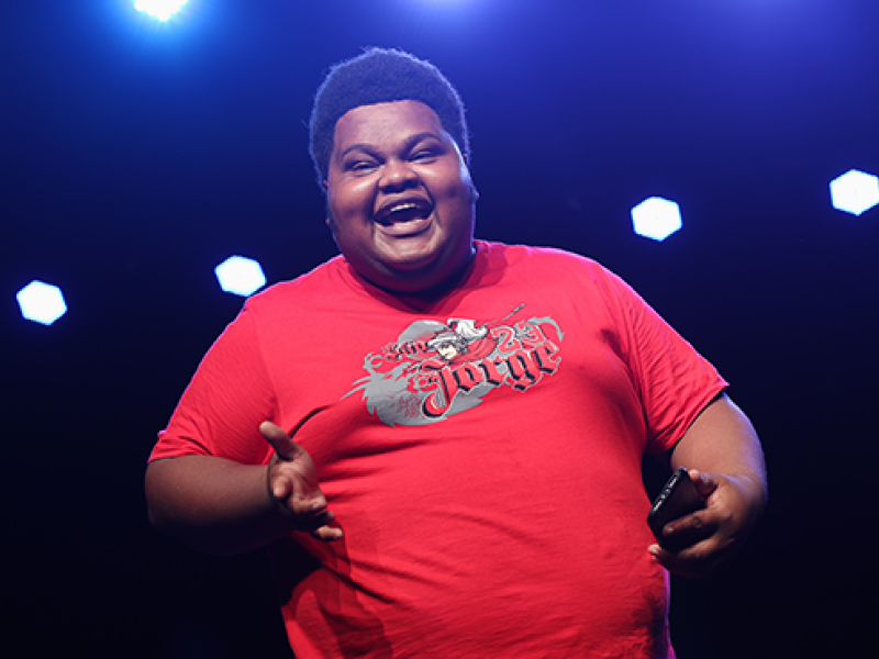 O comediante Big Jaum se apresenta no palco com expressão alegre e sorriso aberto. Vestindo camiseta vermelha e segurando um celular, ele faz um gesto com uma das mãos enquanto interage com o público. A iluminação azul ao fundo cria contraste com a roupa e reforça a atmosfera vibrante do show.