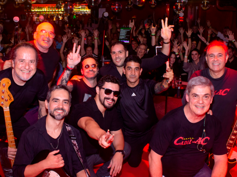 Integrantes da banda Cadillac posam com a plateia ao fundo durante apresentação. Reunidos em frente ao palco, os músicos sorriem e fazem gestos para a câmera, enquanto o público lota a casa de shows com os braços erguidos. A iluminação vermelha e o ambiente festivo destacam a energia do espetáculo.