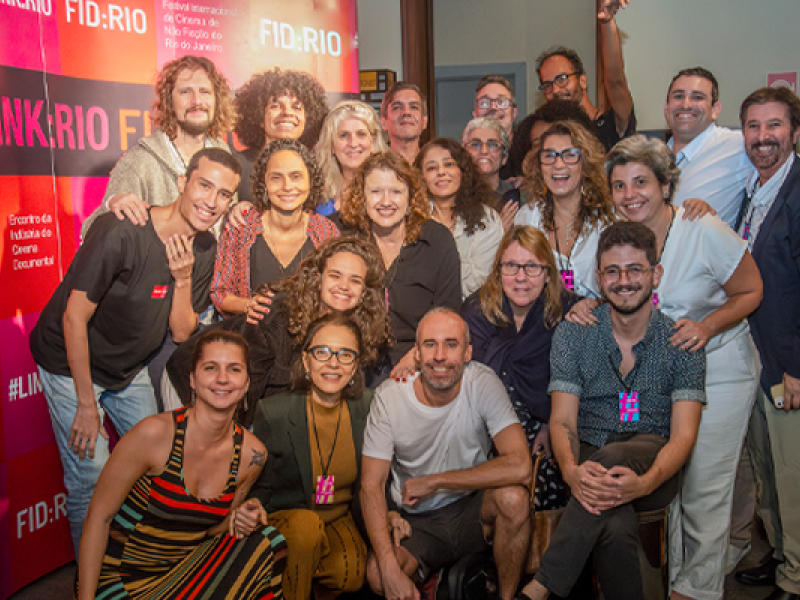 Integrantes da equipe do FID:RIO (Festival Internacional de Cinema de Não Ficção do Rio de Janeiro) posam para fotografia coletiva. Reunidos diante do painel de divulgação do festival, cerca de vinte pessoas sorriem para a câmera em clima de celebração. Ao fundo, a identidade visual do evento reforça o contexto cinematográfico e colaborativo da imagem.