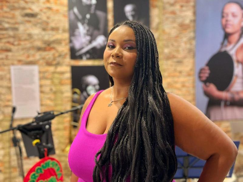 Retrato da cantora Joyce Bella em espaço cultural. Em primeiro plano, ela posa para a câmera com vestido rosa vibrante, cabelos longos e uma das mãos apoiada na cintura. Ao fundo, uma parede de tijolos aparentes exibe fotografias em exposição e um microfone em pedestal, reforçando o contexto musical da imagem.