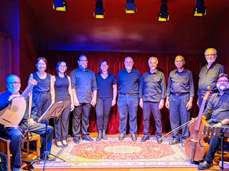 Integrantes do Coro Madrigal da ACC posam em fotografia. O grupo está alinhado diante de uma cortina vermelha, vestindo trajes pretos. Nas extremidades, dois músicos seguram instrumentos de cordas antigos, um alaúde e uma viola da gamba. A iluminação cênica em tons quentes destaca os participantes e o tapete ornamentado que compõe o cenário, criando atmosfera de concerto de música vocal e de câmara.