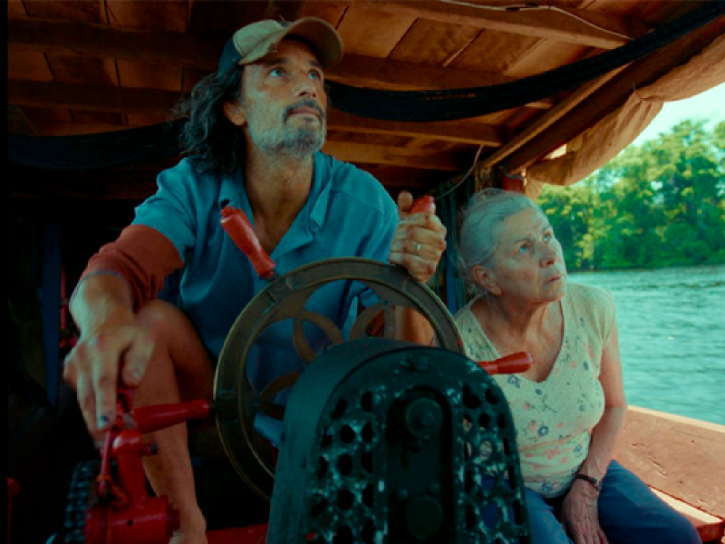 Cena do filme “O Último Azul”, com os atores Rodrigo Santoro e Denise Weinberg. Em uma embarcação sobre um rio, Rodrigo Santoro, de boné e camisa azul, segura o leme enquanto observa algo à frente. Ao seu lado, Denise Weinberg acompanha a mesma direção com expressão atenta. Ao fundo, aparecem a água e a vegetação ribeirinha, reforçando a atmosfera de viagem pelos rios da Amazônia.