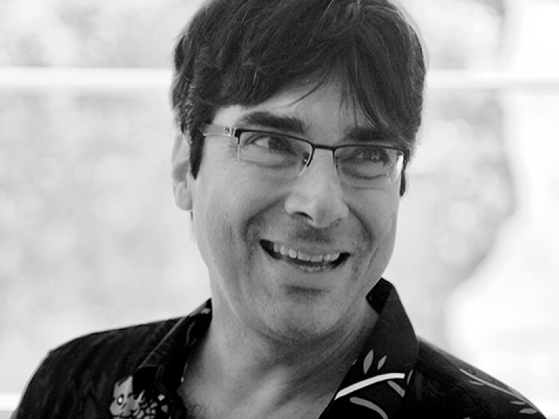 Retrato em preto e branco do poeta Alberto Pucheu. Ele aparece em primeiro plano, usando óculos e camisa escura estampada, com um sorriso espontâneo enquanto olha para fora do enquadramento. O fundo desfocado e a iluminação suave destacam sua expressão acolhedora e a atmosfera descontraída da fotografia.