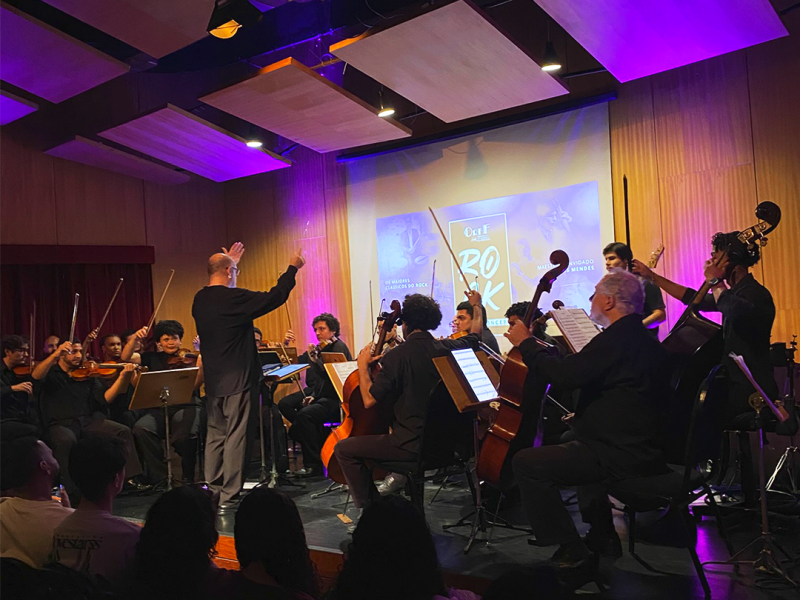 Apresentação da Orquestra Filarmônica Fluminense em palco iluminado por luzes em tons de roxo e azul. Ao centro, o maestro rege os músicos com os braços erguidos. A orquestra é formada por instrumentistas de cordas, incluindo violinos, violoncelos e contrabaixos, dispostos diante do público. Ao fundo, uma tela de projeção exibe a identidade visual do concerto. O ambiente destaca a execução coletiva e a atmosfera solene da apresentação.