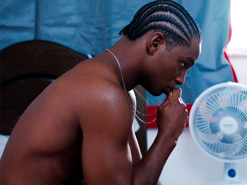 Cena de filme mostra um homem negro sem camisa, usando bermuda vermelha, sentado à beira de uma cama. Com a mão próxima ao rosto e o olhar voltado para baixo, ele aparece em atitude reflexiva. Ao lado, um pequeno ventilador está apoiado sobre uma cadeira plástica branca, diante de uma cortina azul. A composição cria uma atmosfera íntima e contemplativa.