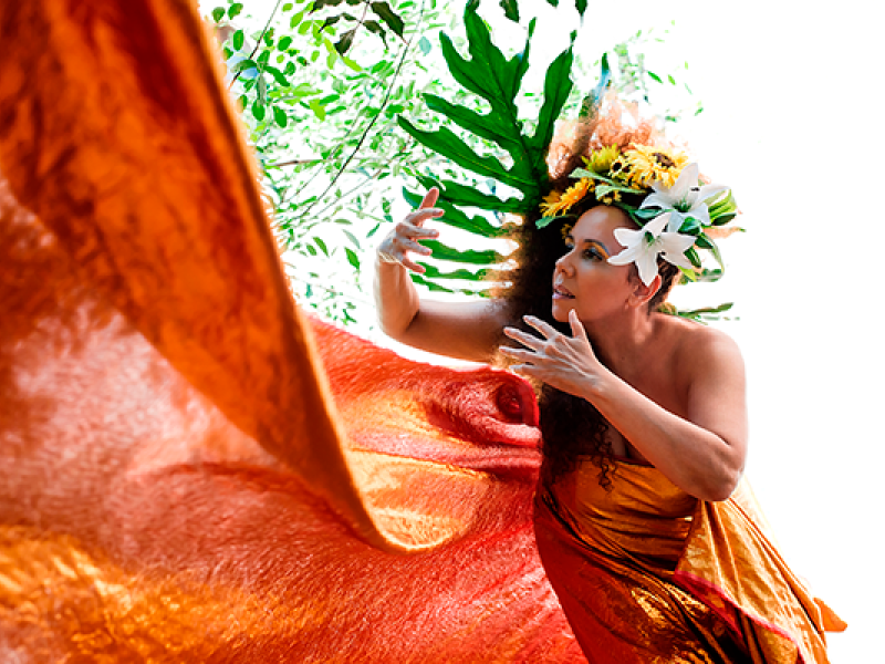 Retrato da cantora Zilá Lima em cenário cercado por elementos da natureza. Com vestido em tons de laranja e adorno floral na cabeça, ela faz gestos expressivos com as mãos enquanto olha para a frente. Grandes folhas verdes e um tecido alaranjado em movimento compõem a cena, criando uma atmosfera vibrante e inspirada na natureza.