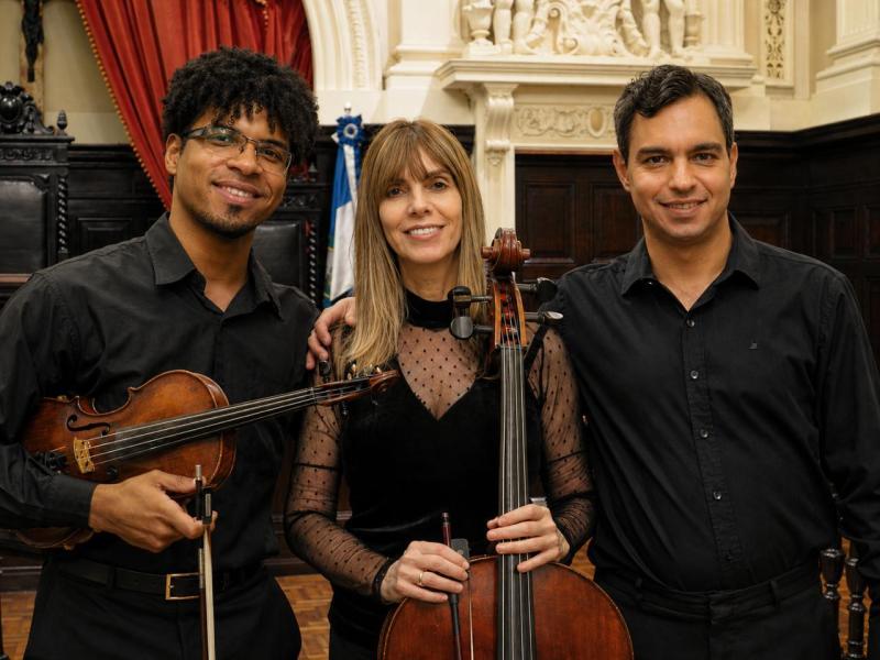 Integrantes do Trio Movimento Musical posam lado a lado em um salão de arquitetura clássica. À esquerda, o violinista Luís Felipe Oliveira segura um violino e o arco. Ao centro, a violoncelista Denise Emmer segura um violoncelo apoiado à frente do corpo. À direita, o pianista João Paulo Romeo sorri para a câmera. Os três vestem roupas pretas e têm ao fundo detalhes em madeira escura, cortinas vermelhas e elementos ornamentados do ambiente.