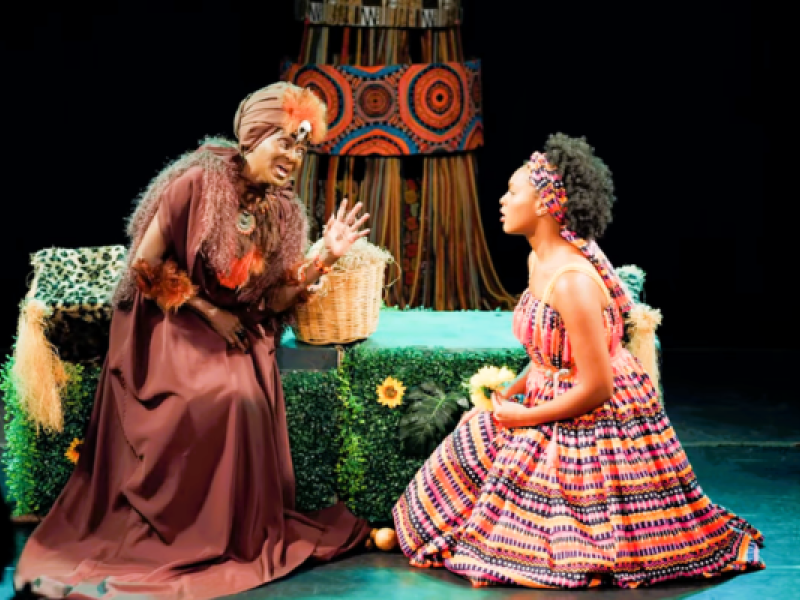 Cena do espetáculo A Bela Adormecida. Em um palco com fundo escuro, uma senhora negra ajoelhada veste longo traje marrom com adornos em tons terrosos, turbante e maquiagem cênica. Ela gesticula enquanto fala com uma jovem negra sentada sobre as pernas à sua frente, que usa vestido amplo e estampado em cores vibrantes, com faixa colorida nos cabelos, e a observa atentamente. Ao lado da senhora há um cesto de vime. Ao fundo, um elemento cenográfico com estampas geométricas africanas completa a composição.