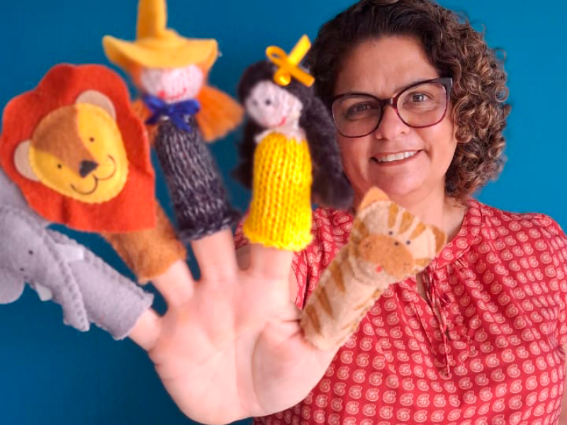 A escritora Luciana Belém sorri para a câmera enquanto exibe dedoches de personagens infantis colocados nos dedos de uma das mãos: um elefante, um leão, um menino, uma menina e um tigre. Ao fundo, um painel azul destaca a cena. A escritora usa óculos, blusa estampada em tons de vermelho e mantém a mão estendida em direção à câmera, aproximando os dedoches do primeiro plano.