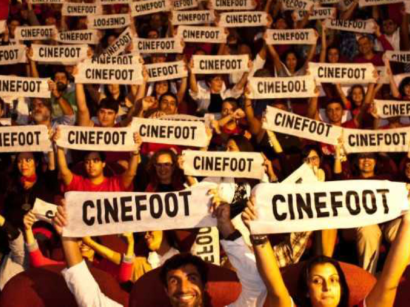 Plateia lotada em um cinema ergue dezenas de faixas brancas com a palavra "CINEFOOT" impressa em letras pretas. Pessoas de diferentes idades sorriem e levantam os cartazes acima da cabeça, criando um efeito coletivo que ocupa quase toda a imagem e transmite entusiasmo e celebração em torno do festival.