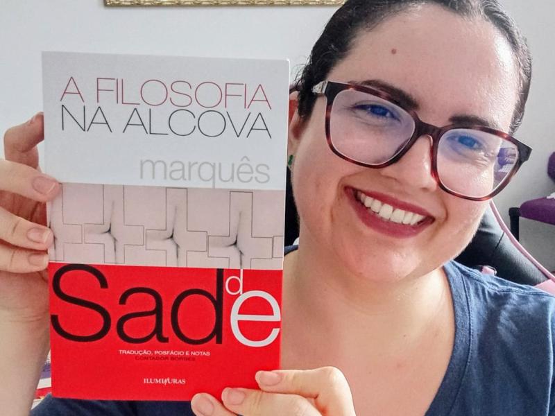 A pesquisadora Nicole Ayres sorri para a câmera enquanto segura o livro A Filosofia na Alcova, do Marquês de Sade, com a capa voltada para a frente. Ela usa óculos de armação escura e blusa azul. O livro, em tons de branco, cinza e vermelho, ocupa boa parte da imagem e destaca o título e o nome do autor.