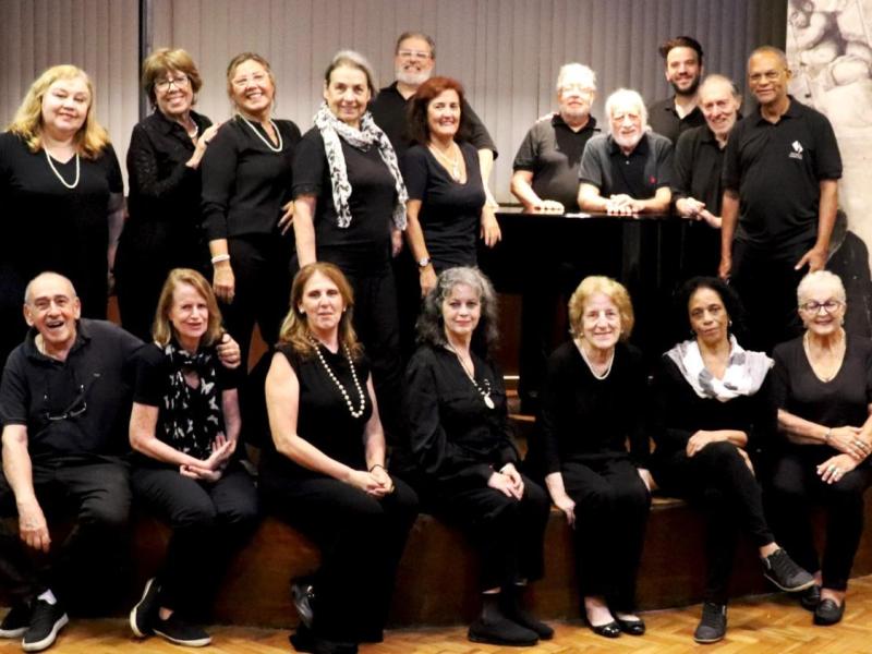 Integrantes do Coro Tu Voz, Mi Voz, da Associação de Canto Coral, posam reunidos em um palco. O grupo é formado por homens e mulheres de diferentes idades, distribuídos entre pessoas sentadas à frente e outras em pé ao fundo, todas vestindo roupas predominantemente pretas. Ao centro, um piano reúne parte dos integrantes, enquanto o piso de madeira e as cortinas ao fundo compõem o ambiente da apresentação.
