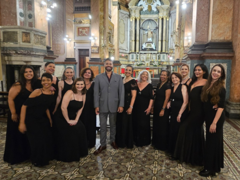 Integrantes do Coro Lírico Feminino (ACC) posam ao lado do maestro Cláudio Ávila no interior de uma igreja. O maestro, de terno cinza, está ao centro, cercado pelas cantoras, que vestem longos vestidos pretos. Ao fundo, o altar ornamentado, colunas de mármore e lustres iluminados destacam a arquitetura do templo.