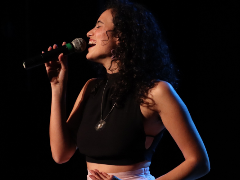 A cantautora, arranjadora vocal e professora de canto Katarina Assef canta ao microfone em um palco com fundo escuro. Vista de perfil, ela sorri com os olhos fechados, segura o microfone com uma das mãos e apoia a outra sobre o abdômen. Usa blusa preta sem mangas, calça clara e um colar com pingente prateado. A iluminação destaca seu rosto e sua expressão durante a apresentação.