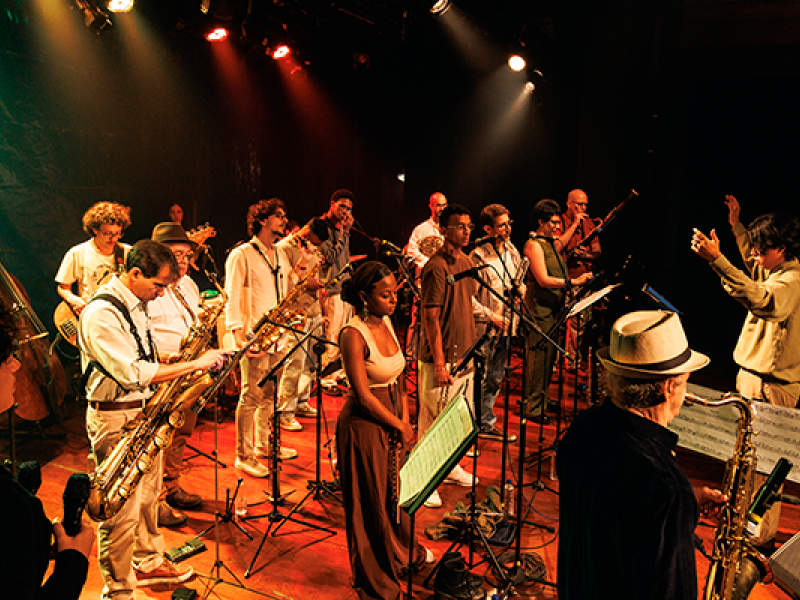 Integrantes da Orquestra Mil Tons se apresentam em um palco iluminado por refletores. O grupo reúne músicos, cantores e instrumentistas distribuídos entre microfones e estantes de partitura. À direita, o maestro rege a apresentação com os braços erguidos, enquanto, ao fundo, diferentes instrumentos de sopro, cordas e percussão compõem a cena. A iluminação em tons quentes reforça o clima vibrante do espetáculo.
