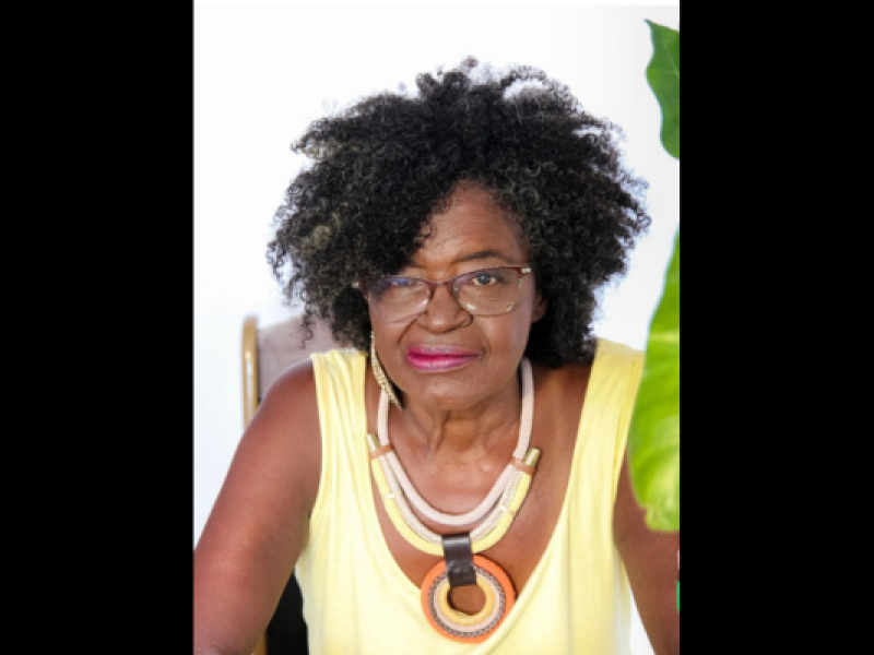 Retrato da escritora e poeta Miriam Alves. Ela olha diretamente para a câmera, com expressão serena e leve sorriso. Tem pele negra, cabelos crespos volumosos com fios grisalhos, usa óculos de armação discreta, blusa amarela sem mangas e um colar de grandes argolas em tons de bege, marrom e laranja. Ao fundo, há uma parede clara e, à direita da imagem, parte de uma folha verde aparece em primeiro plano.