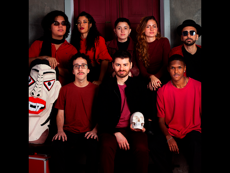 Integrantes da banda autoral Magnífica Máquina Maldita posam em dois níveis diante de uma porta vermelha. O grupo reúne oito jovens com roupas em tons de vermelho, vinho e preto. Em primeiro plano, um dos integrantes segura uma caveira cenográfica, enquanto, à esquerda, uma grande máscara estilizada com traços geométricos completa a composição. Alguns integrantes usam óculos escuros, reforçando a estética contemporânea e teatral da imagem.