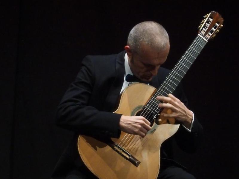 O violonista italiano Fabio Montomoli toca violão clássico em um palco de fundo escuro. Vestindo terno preto, camisa branca e gravata borboleta, mantém a cabeça inclinada em direção ao instrumento enquanto as duas mãos executam a música sobre as cordas e o braço do violão. A iluminação destaca o músico e o instrumento, reforçando a atmosfera intimista da apresentação.