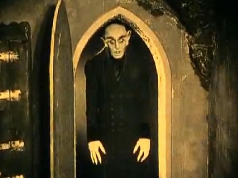 Cena do filme Nosferatu. Um homem de aparência cadavérica, com cabeça raspada, orelhas pontiagudas e dedos longos, surge parado à entrada de uma porta em arco, envolto por sombras. Vestido com roupas escuras, mantém a cabeça levemente inclinada e os braços estendidos ao lado do corpo. A iluminação em tons amarelados e o contraste intenso entre luz e escuridão reforçam a atmosfera sombria da cena.