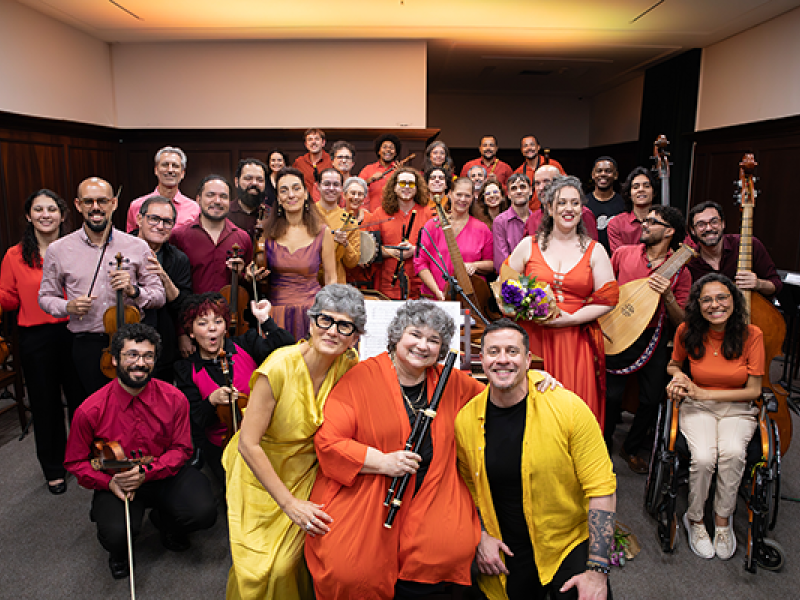 Integrantes da Orquestra Barroca da UNIRIO posam reunidos em uma sala de concertos. O grupo é formado por músicos de diferentes idades, distribuídos em várias fileiras, alguns segurando instrumentos de cordas, sopro e dedilhados. Em primeiro plano, três integrantes vestindo roupas em tons de amarelo e laranja ocupam o centro da composição, enquanto, ao fundo, os demais sorriem para a câmera. A imagem destaca a diversidade do conjunto e a atmosfera de celebração entre os músicos.