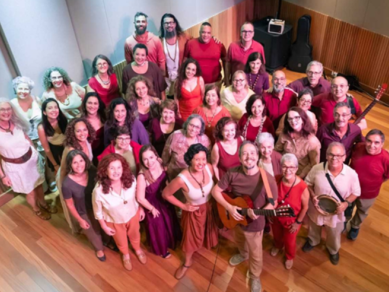 Integrantes do coro Ordizão posam para uma fotografia em grupo em uma sala de ensaio com piso de madeira. Cerca de quarenta participantes, de diferentes idades, sorriem para a câmera, distribuídos em várias fileiras e fotografados de um ângulo alto. As roupas em tons de vermelho, vinho, rosa, marrom e bege criam uma composição harmoniosa e acolhedora. Em primeiro plano, um integrante segura um violão, enquanto outro segura um pandeiro. Ao fundo, encostados na parede, aparecem um violão e um amplificador.