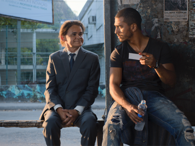 Cena do filme Papagaios. Em um ponto de ônibus, dois homens estão sentados lado a lado. À esquerda, um homem de baixa estatura veste terno cinza, camisa branca e gravata, mantendo as mãos apoiadas sobre o colo enquanto sorri discretamente para o homem ao lado. À direita, um homem de camiseta escura e calça jeans rasgada segura um pequeno cartão branco e uma garrafa de água, retribuindo o olhar do companheiro. Ao fundo, a paisagem urbana é iluminada pela luz dourada do fim da tarde.