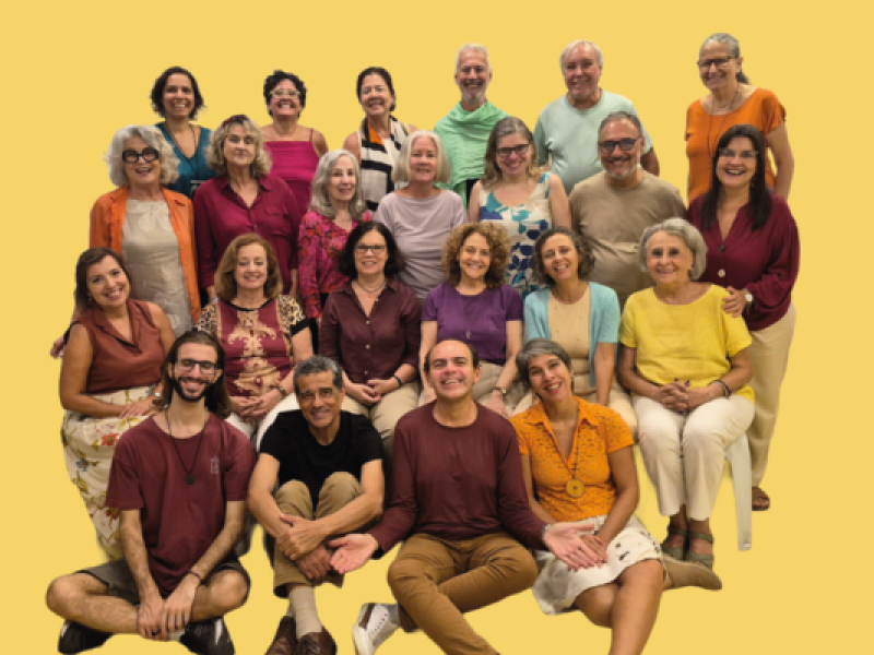 O Coral Pedra da Urca reúne cerca de vinte e cinco integrantes em um retrato coletivo sobre fundo amarelo-claro. Os participantes, distribuídos em quatro fileiras, sorriem para a câmera e vestem roupas em cores variadas, predominando tons quentes e coloridos. Alguns estão em pé, outros sentados e três aparecem acomodados no chão à frente do grupo, compondo uma fotografia descontraída.