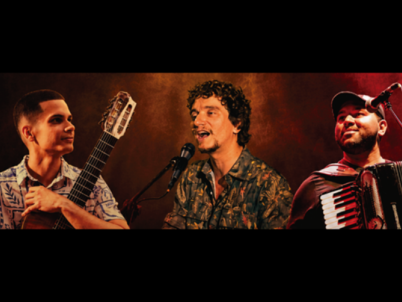 Os integrantes do trio Três em Travessia aparecem lado a lado sobre um fundo em tons de marrom e laranja. À esquerda, o violonista de sete cordas Igor Souza segura o instrumento e olha para o centro. Ao centro, o cantor e pianista Gabriel Ruiz canta ao microfone. À direita, o sanfoneiro e cantor Rafael Meninão toca sanfona enquanto olha para cima com um leve sorriso.