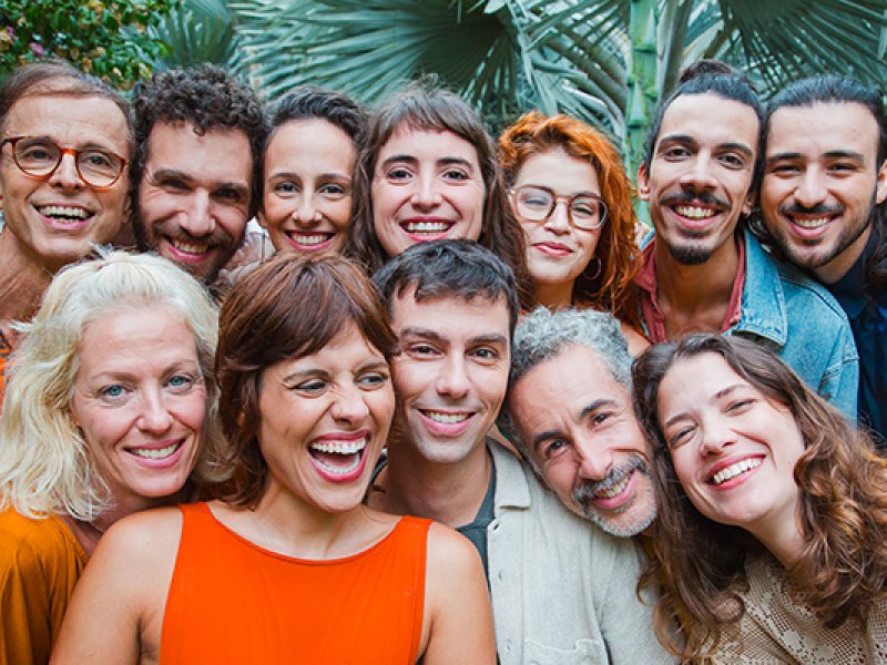 Integrantes do grupo vocal Vozerê sorriem reunidos para uma fotografia em close. O grupo é formado por doze pessoas de diferentes idades, posicionadas bem próximas umas das outras diante de um jardim com folhagens ao fundo. As expressões alegres e a variedade de sorrisos, olhares e gestos transmitem um clima de amizade, acolhimento e celebração.