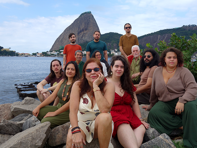 Integrantes do Coral Altivoz posam reunidos sobre pedras à beira da água, tendo ao fundo o Pão de Açúcar e a paisagem da Baía de Guanabara. O grupo é formado por pessoas de diferentes idades, distribuídas em vários níveis, algumas sentadas e outras em pé. Vestindo roupas em tons terrosos, verdes, vermelhos e bege, os integrantes sorriem ou mantêm expressões serenas, compondo um retrato coletivo em ambiente ao ar livre.