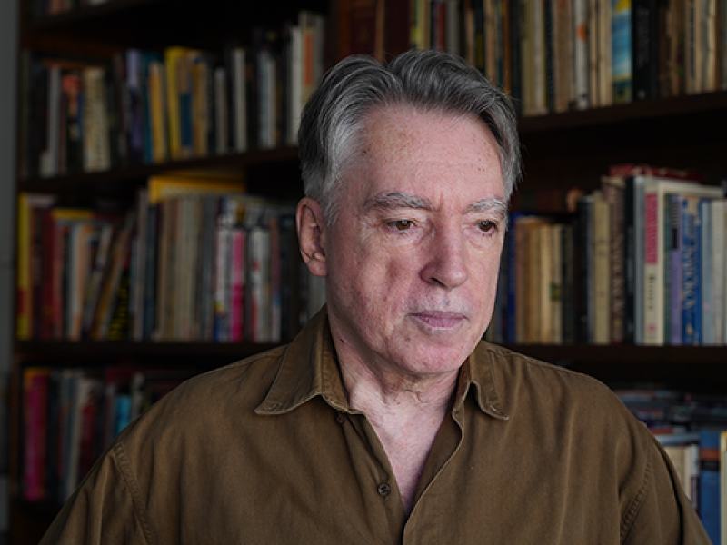O poeta Braulio Tavares é retratado diante de uma estante repleta de livros. Com cabelos grisalhos penteados para trás e camisa marrom, mantém o olhar voltado levemente para baixo, com expressão serena e reflexiva. O fundo desfocado destaca as prateleiras repletas de livros, reforçando o ambiente de leitura e criação literária.