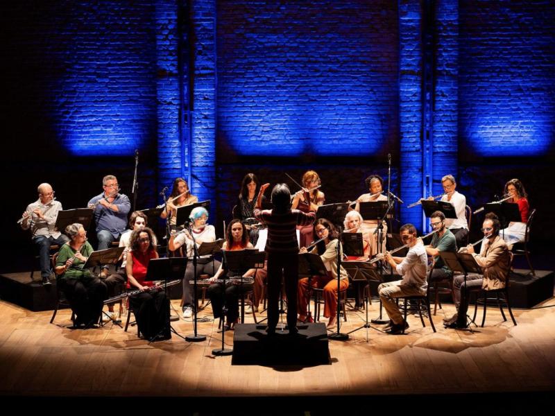 Apresentação da Orquestra de Flautas da Casa do Choro. No palco, cerca de vinte músicos estão dispostos em semicírculo, com partituras à frente, tocando flautas de diferentes tamanhos. Ao centro, de costas para a câmera, o regente conduz o conjunto sobre um pequeno tablado. Ao fundo, uma parede de tijolos é destacada por iluminação azul.