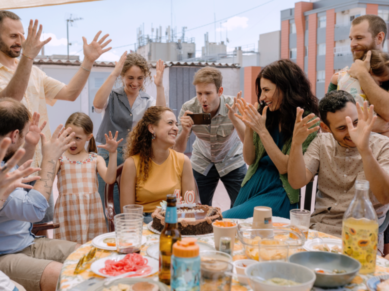 Still de um dos filmes da mostra de cinema espanhol. Um grupo de adultos e uma criança está reunido ao redor de uma mesa durante uma comemoração. Ao centro, uma mulher sorri diante de um bolo com velas que formam o número 40, enquanto as pessoas ao redor levantam as mãos em gestos animados. A mesa está repleta de pratos, copos e bebidas, e a cena acontece em uma área externa cercada por edifícios.