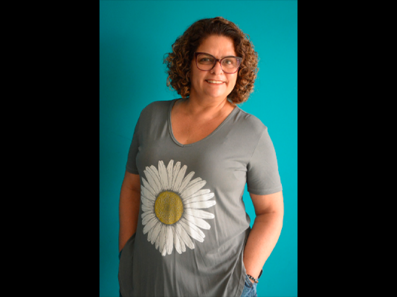 Retrato da escritora Luciana Belém. Ela aparece em pé, sorrindo para a câmera, com cabelos castanhos cacheados e óculos. Veste blusa cinza com a estampa de uma grande margarida branca e mantém as mãos nos bolsos. Ao fundo, uma parede azul-turquesa contrasta com a roupa.